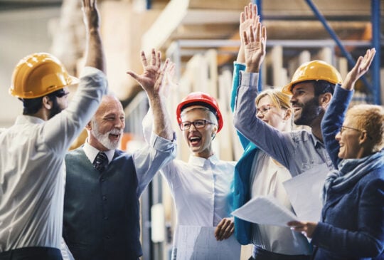 Closeup side view of mixed age people of industrial design department at a factory. They are gathered at a desk and celebrating successful job by giving a high five.